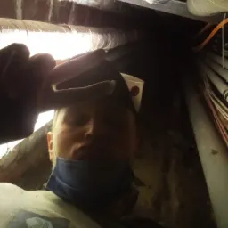 Mężczyzna w roboczym ubraniu z logiem firmy, prawdopodobnie w trakcie pracy w ciasnej przestrzeni pod stropem, otoczony rurami i przewodami, z kubkiem w kieszeni czapki.