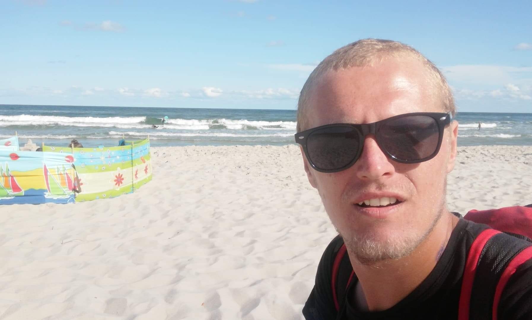 Mężczyzna w okularach przeciwsłonecznych robiący selfie na piaszczystej plaży z widokiem na morze i parawan plażowy w tle.