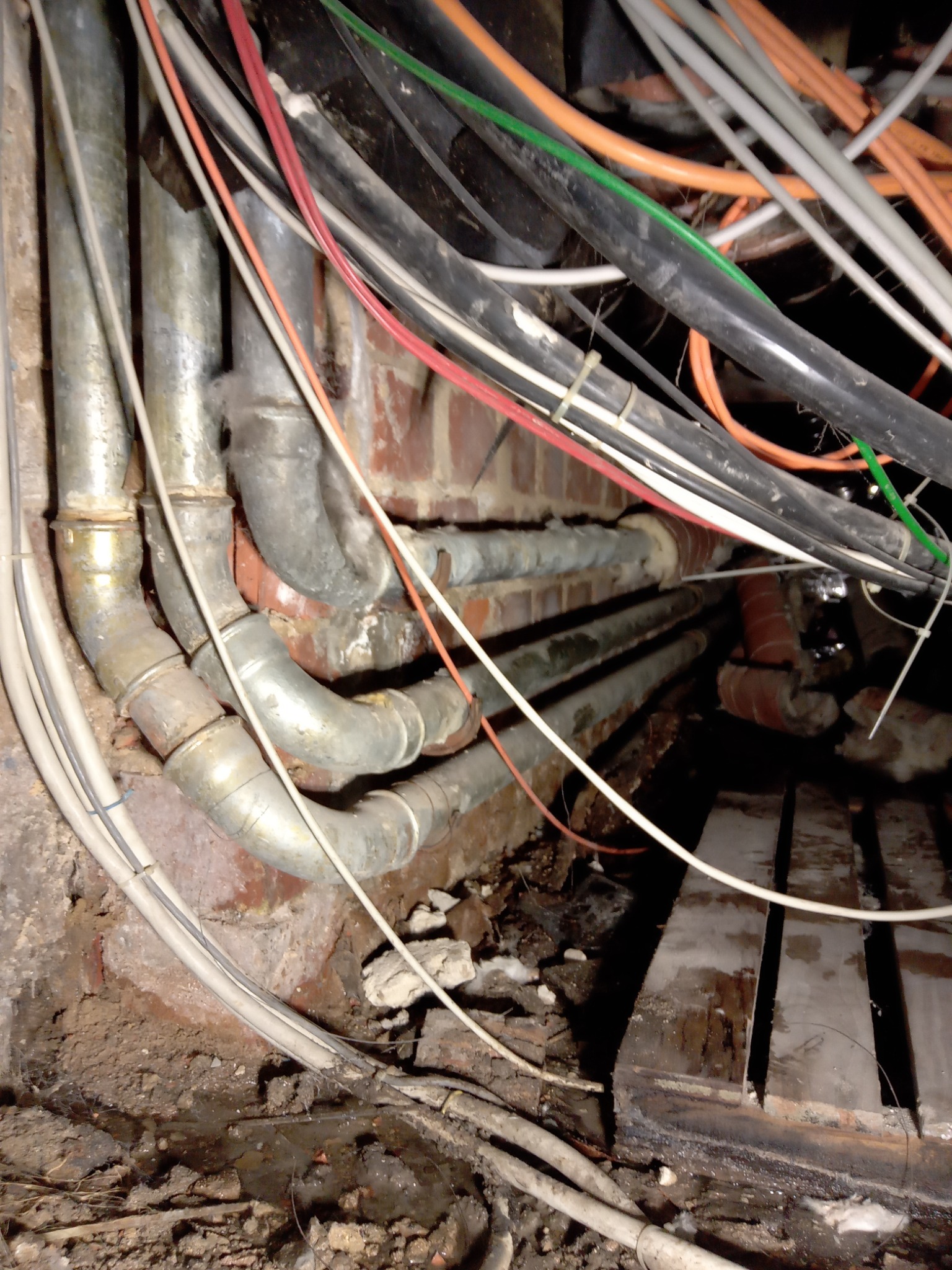 Złożona sieć rur i kabli w przestrzeni pod podłogą, widoczne fragmenty ceglanej ściany i drewnianej konstrukcji podłogi, oświetlenie słabe, przewody w różnych kolorach spięte opaskami.