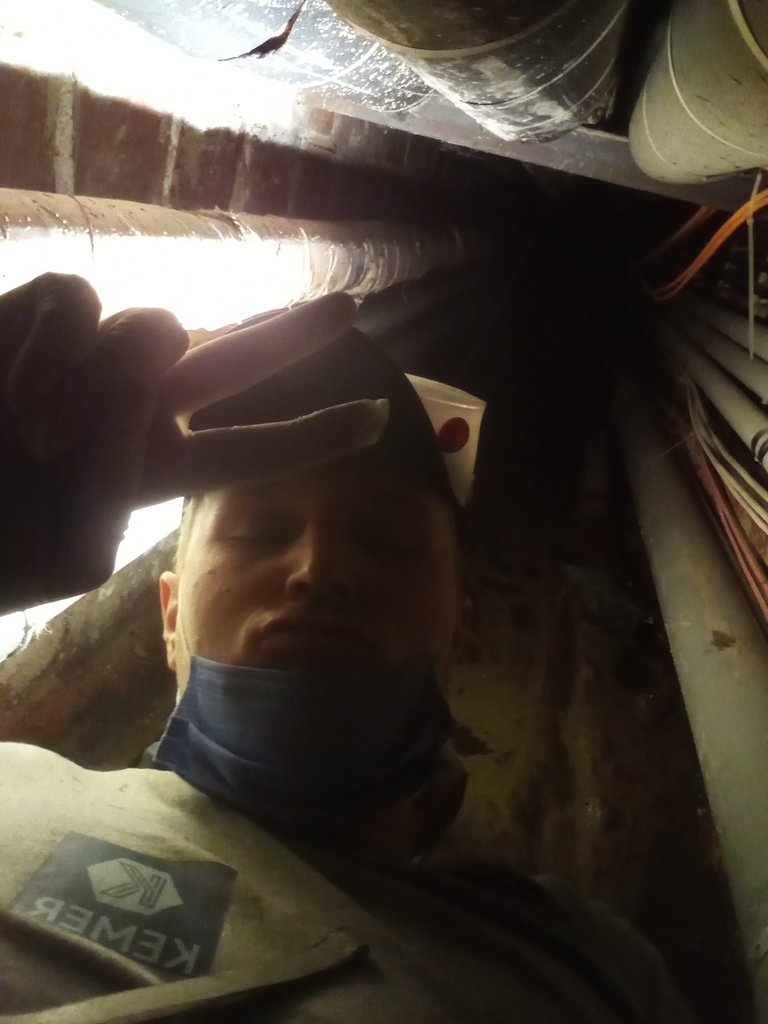 Mężczyzna w roboczym ubraniu z logiem firmy, prawdopodobnie w trakcie pracy w ciasnej przestrzeni pod stropem, otoczony rurami i przewodami, z kubkiem w kieszeni czapki.