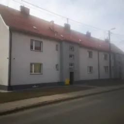 Trzysegmentowy budynek mieszkalny z czerwonym dachem, widoczny z ulicy w pochmurny dzień.