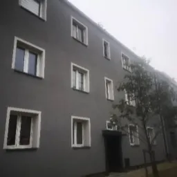 Szary budynek wielorodzinny po ociepleniu w Zabrzu, widoczne białe ramy okienne i ciemne drzwi wejściowe, ujęcie w pochmurny dzień.