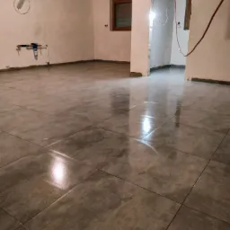 Ułożona, błyszcząca, szara podłoga z płytek w surowym wnętrzu, widoczne instalacje wodne na ścianie i otwór drzwiowy do kolejnego pomieszczenia.