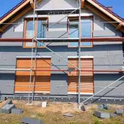 Dom jednorodzinny w trakcie ocieplania, widoczne szare płyty izolacyjne na elewacji, rusztowanie z metalowych rur, okna z brązowymi roletami, fragment dachu z brązową dachówką, na trawie leżą...