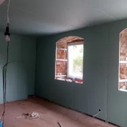 Montaż płyt kartonowo-gipsowych na ścianach i suficie w pomieszczeniu z oknami o zaokrąglonych łukach, widoczne profile mocujące, instalacja elektryczna wystająca ze ściany, surowy stan podłogi.