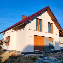 Dom jednorodzinny z jasną elewacją i brązową stolarką okienną, częściowo zakryte rolety zewnętrzne, widoczne materiały budowlane i rusztowanie w trakcie prac wykończeniowych.