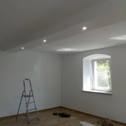 Wnętrze pomalowanego na biało pokoju z drewnopodobną podłogą, widoczna drabina aluminiowa, listwy przypodłogowe, narzędzia i odkurzacz, okno z łukowym nadprożem wpuszczające jasne światło.