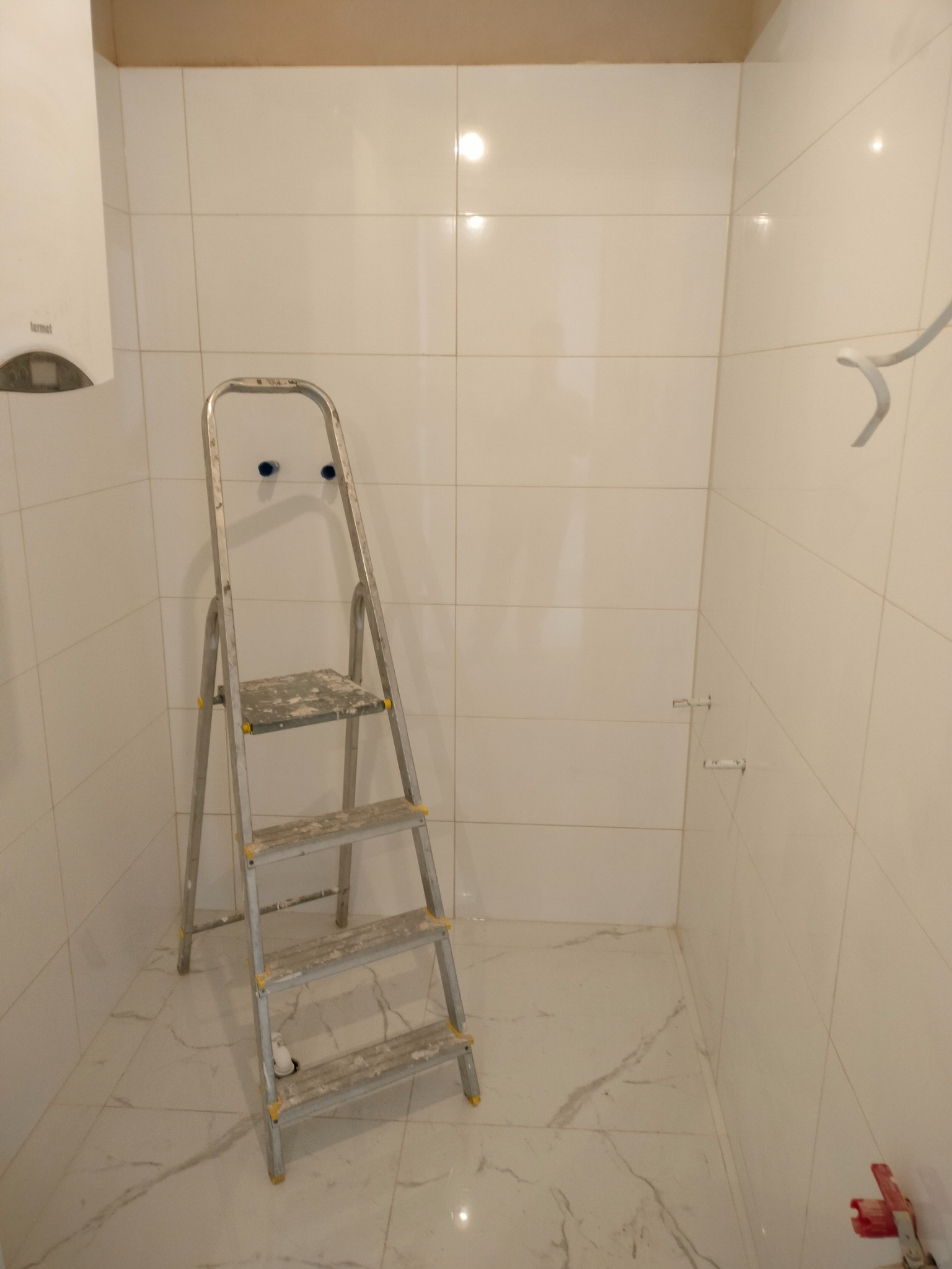 Wnętrze łazienki w trakcie wykańczania, białe płytki na ścianach i podłodze imitujące marmur, drabina malarska z resztkami zaprawy, grzejnik elektryczny na ścianie, widoczne przyłącza hydrauliczne.