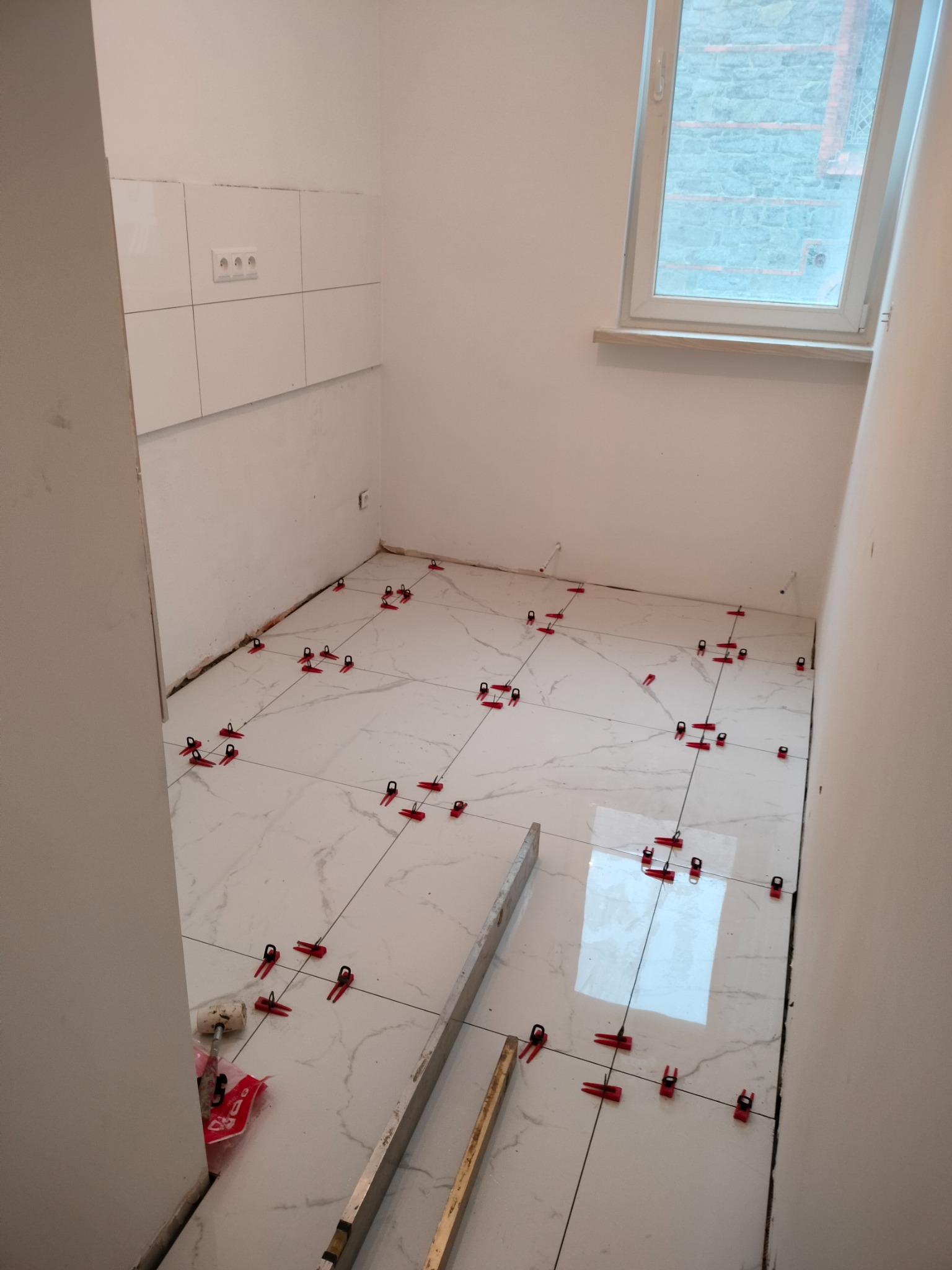 Białe płytki podłogowe imitujące marmur, ułożone w kwadraty z użyciem czerwonych klipsów poziomujących, widoczne narzędzia i poziomica, okno w tle.