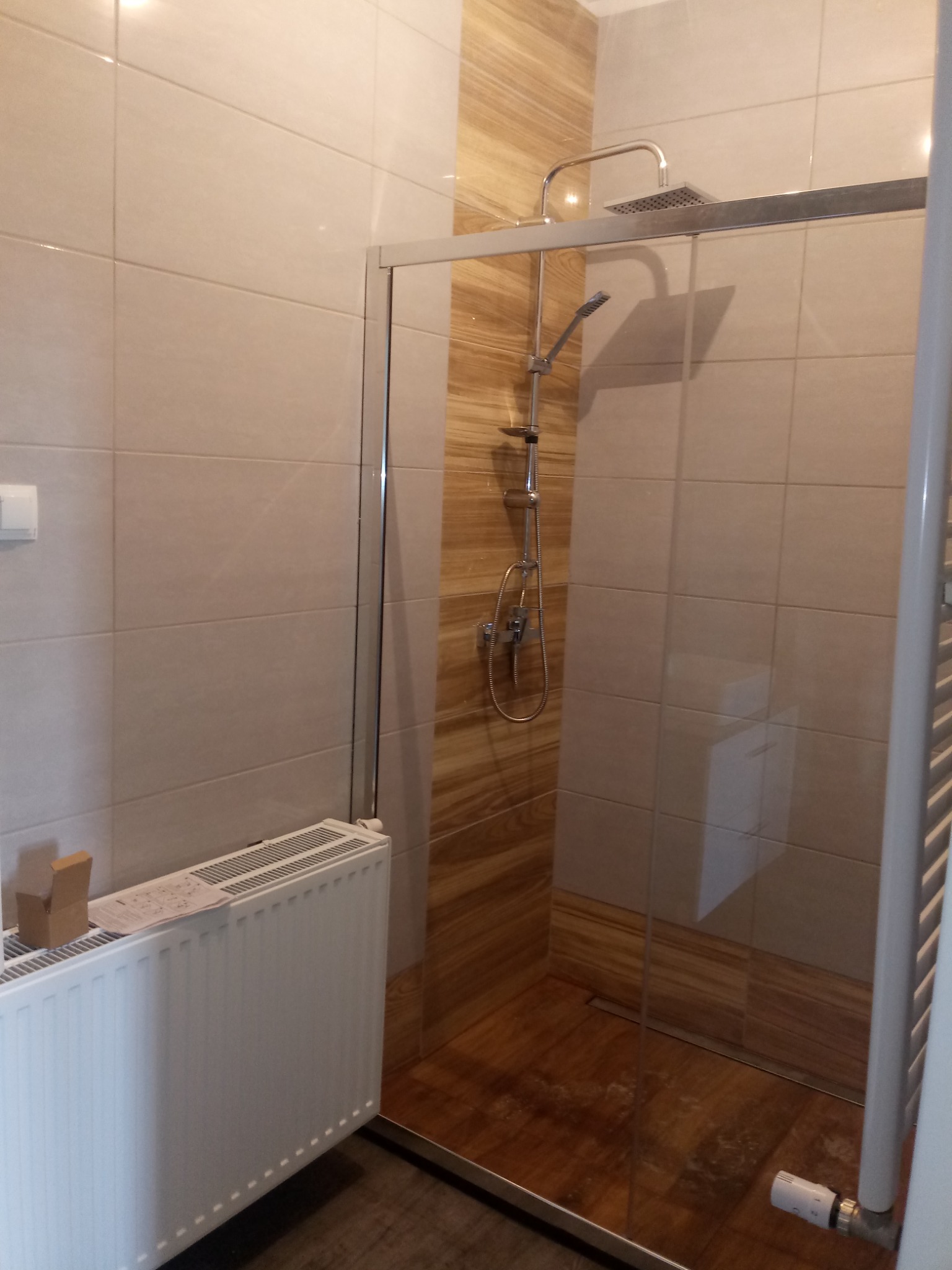Nowoczesna kabina prysznicowa z deszczownicą, obłożona beżowymi i drewnopodobnymi płytkami ceramicznymi, z widocznym grzejnikiem i kartonem na nim.