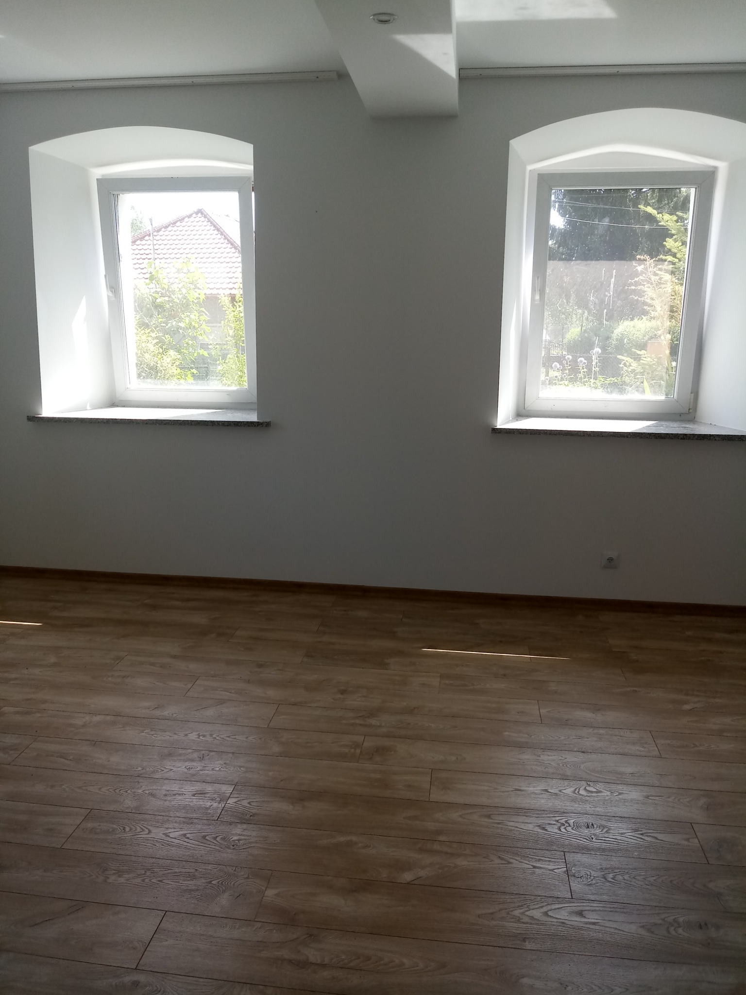 Puste wnętrze z nową podłogą imitującą drewno, świeżo pomalowanymi białymi ścianami i dwoma oknami z zaokrąglonymi górnymi krawędziami wnęk okiennych.