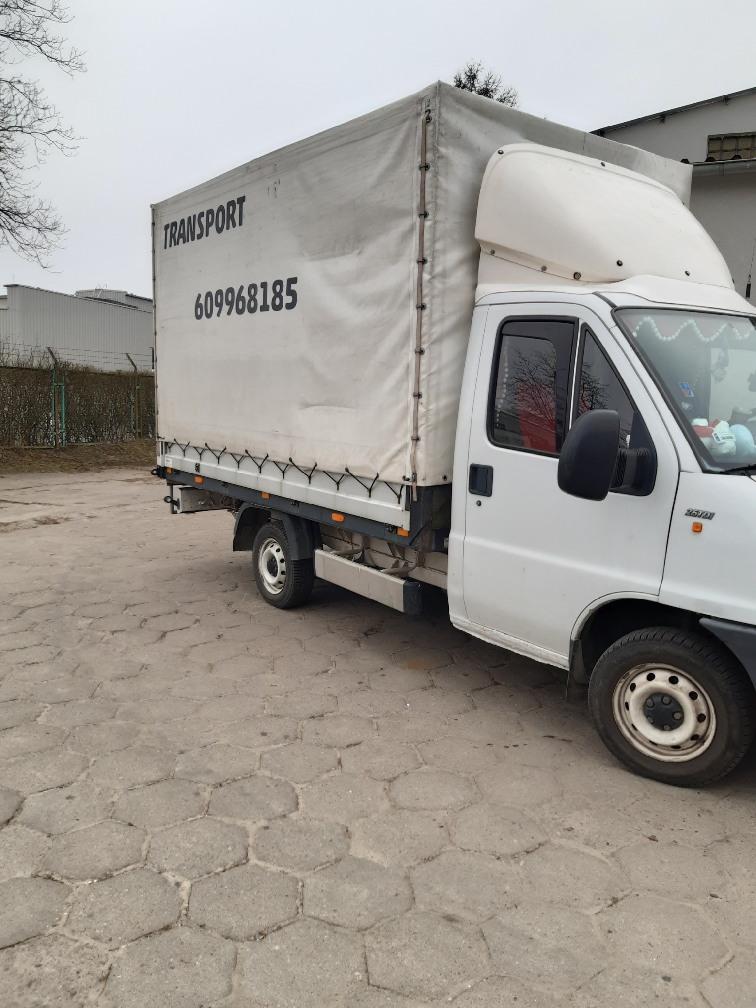 Biały samochód dostawczy z plandeką, na której widnieje napis 'TRANSPORT' i numer telefonu 609968185, zaparkowany na betonowym placu w pochmurny dzień.