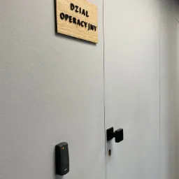 Ściana z szarą teksturą, na której zamontowano drewnianą tabliczkę z napisem 'Dział Operacyjny' w czarnych literach oraz czarny czytnik kart dostępu z diodami LED, obok białych drzwi z czarną klamką.