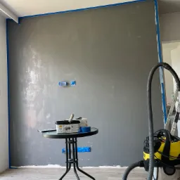 Ściana w trakcie malowania na szaro, zabezpieczona niebieską taśmą malarską, widoczny stolik z farbami i akcesoriami oraz odkurzacz przemysłowy Karcher.