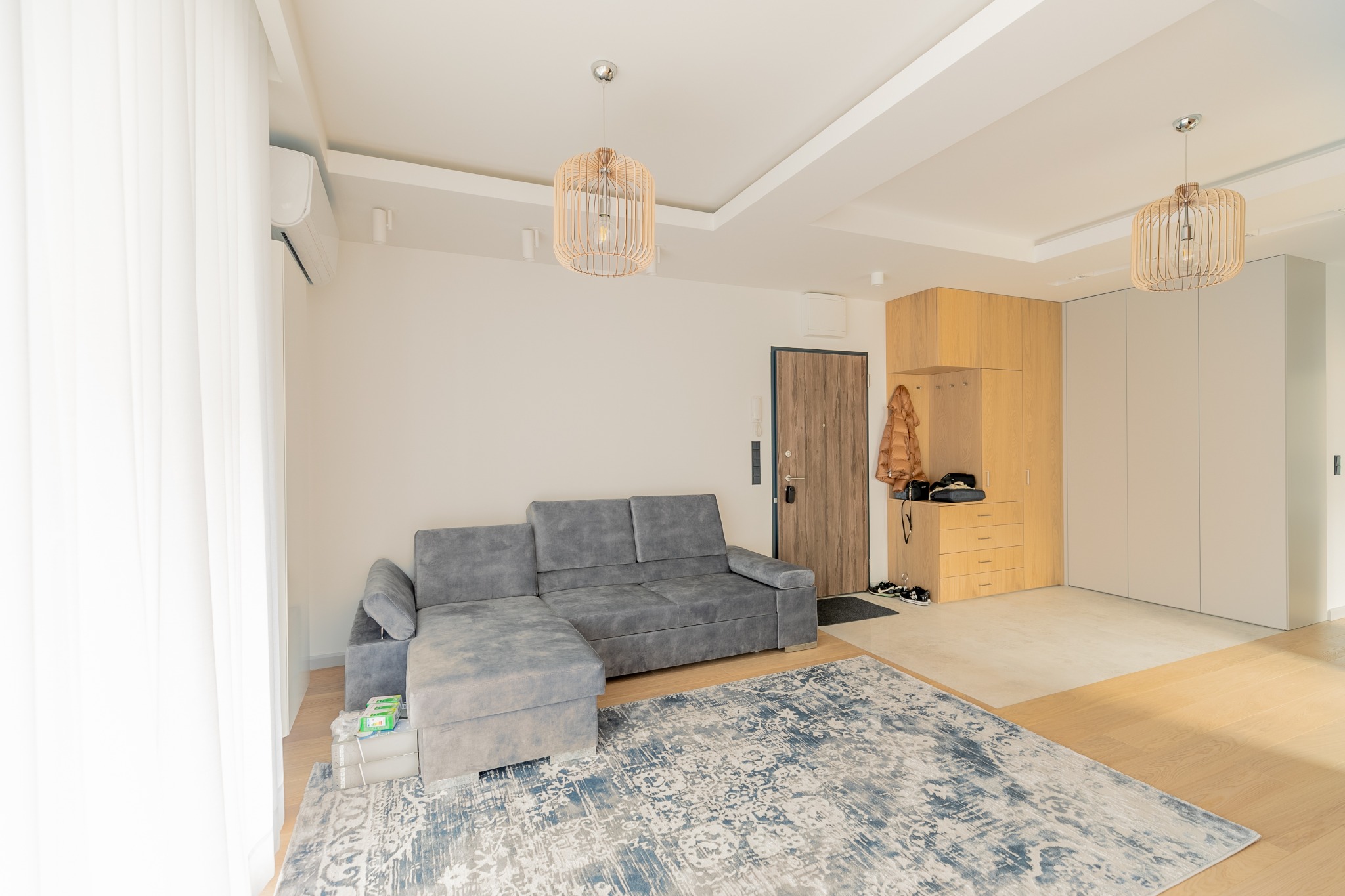 Jasne wnętrze mieszkania z szarą sofą narożną, wzorzystym dywanem w odcieniach błękitu i beżu, drewnianą szafą w zabudowie i ozdobnymi lampami wiszącymi z drewnianymi elementami.