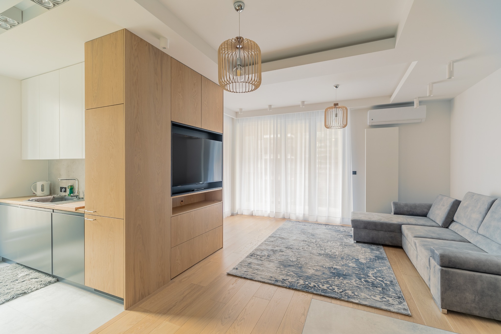 Jasne, nowoczesne wnętrze mieszkania z aneksem kuchennym, drewnianą zabudową z telewizorem, szarą sofą narożną i dywanem z niebieskim wzorem.