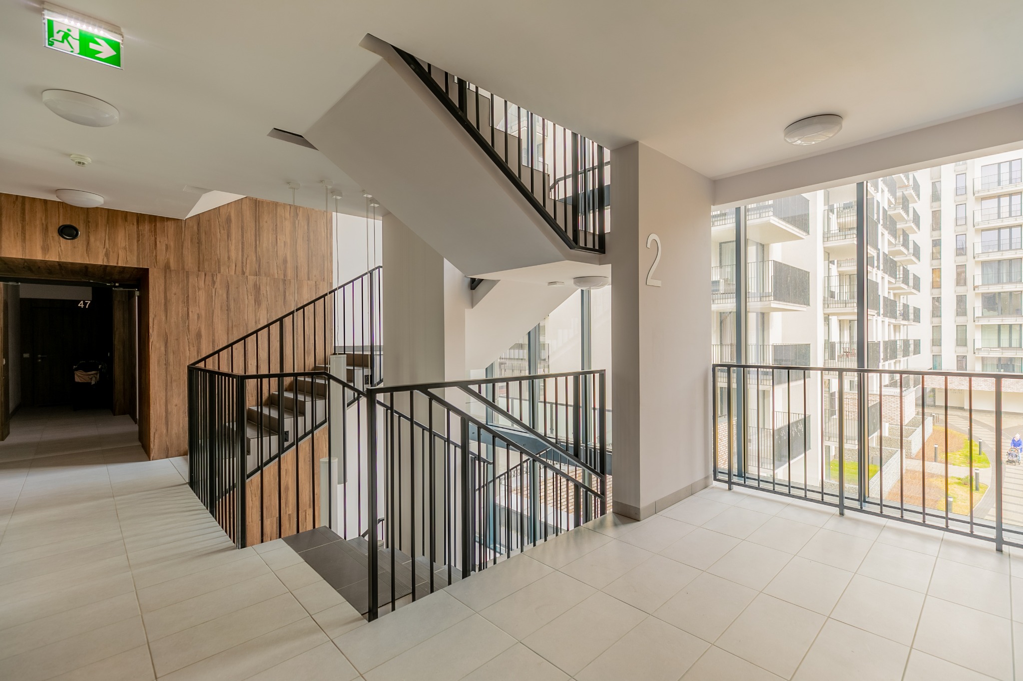 Klatka schodowa w nowoczesnym apartamentowcu z czarnymi balustradami, numerem 2 na ścianie i widokiem na inne budynki przez okno.