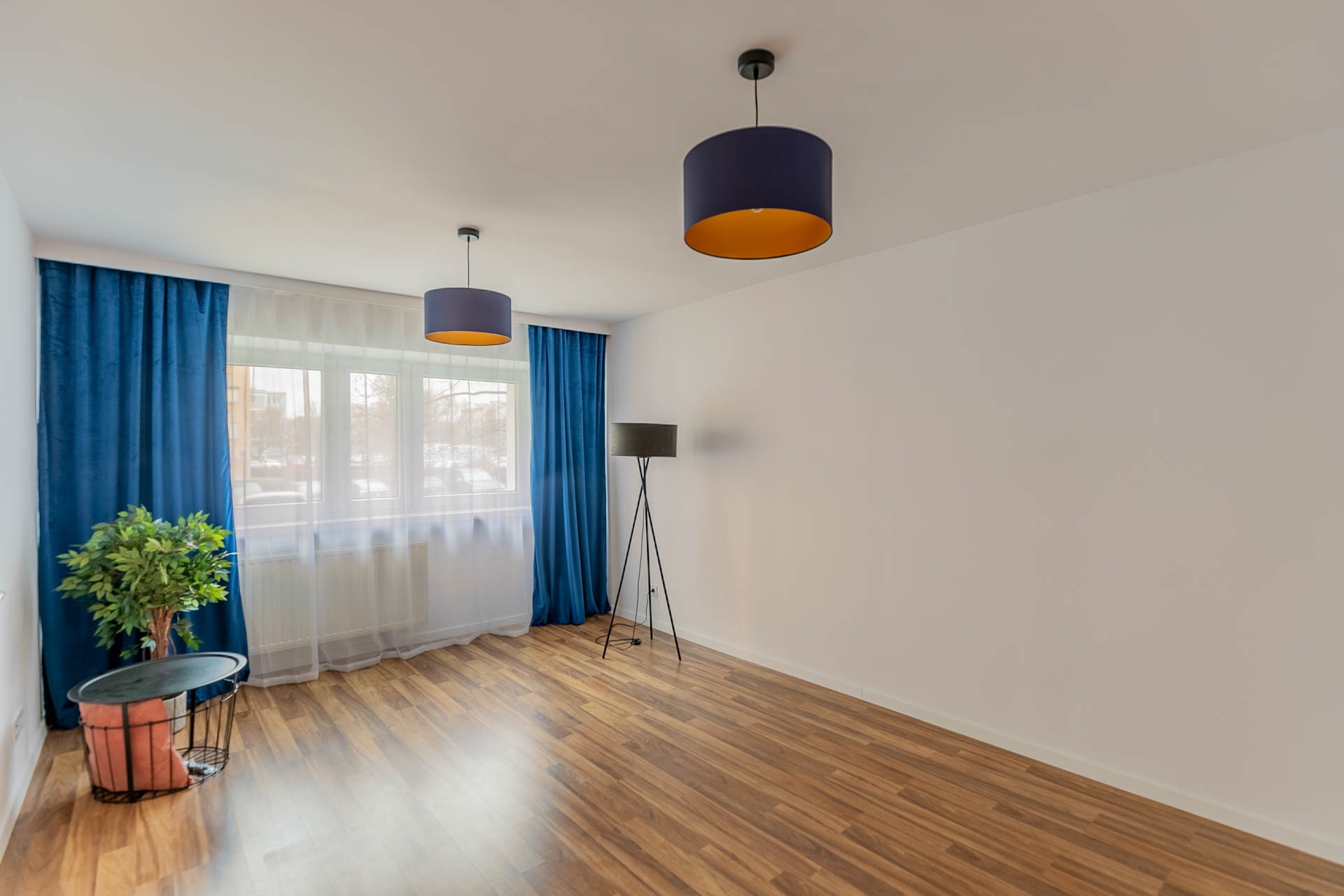 Puste, jasne wnętrze z drewnianą podłogą, niebieskimi zasłonami, lampą stojącą na trójnogu i dwoma wiszącymi lampami z niebieskimi abażurami i złotym wnętrzem, roślina w ozdobnym koszu.