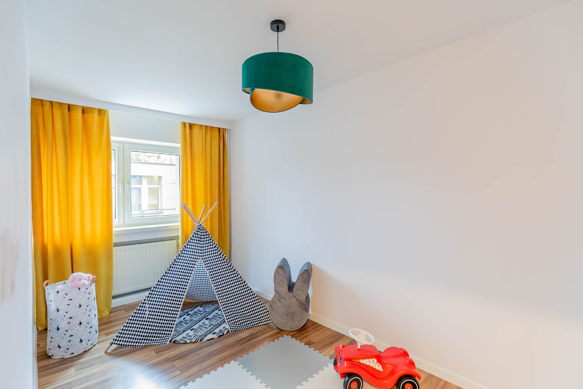 Jasny pokój dziecięcy z żółtymi zasłonami, tipi w czarno-białą kratę, szarym pufem w kształcie królika, czerwonym jeździkiem i zielono-złotą lampą wiszącą.