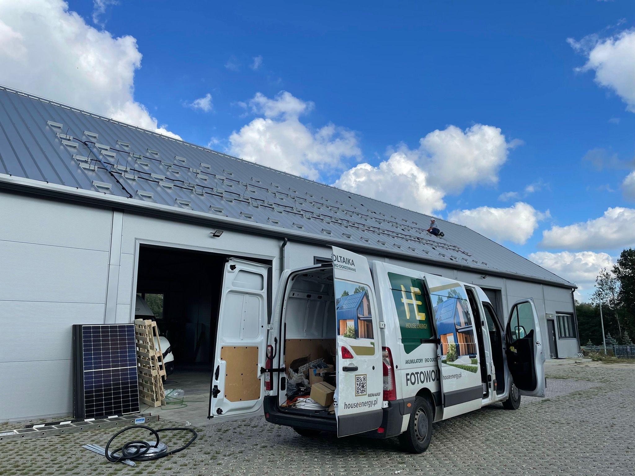 Montaż paneli fotowoltaicznych na dachu z blachy trapezowej. Widoczny bus firmowy z brandingiem, moduły fotowoltaiczne oraz pracownik na dachu podczas instalacji.