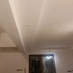 Wnętrze w trakcie prac wykończeniowych: świeżo pomalowany sufit z widocznymi niedoskonałościami i przewodem, ściany przygotowane do malowania, drzwi zabezpieczone folią. Widoczne ślady po taśmie...