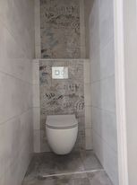 Kompakt WC podwieszany z przyciskiem spłukującym na tle szarej, wzorzystej płytki, obramowany białymi płytkami imitującymi marmur w małej łazience.