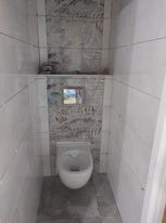 Wąska toaleta podwieszana w małej wnęce, wykończona białymi i szarymi płytkami o wzorze marmuru, widoczny przycisk spłukiwania, surowy minimalizm.