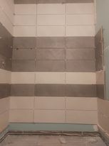 Ściana łazienki w trakcie układania płytek w poziome pasy w odcieniach szarości i beżu, widoczne krzyżyki dystansowe i niedokończone krawędzie.