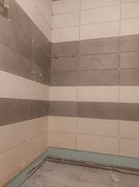 Ściana łazienki w trakcie układania płytek w poziome pasy szaro-białe, widoczna dolna krawędź z zabezpieczeniem przed zabrudzeniem, niedokończone fugowanie.