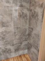 Szare płytki imitujące beton w rogu łazienki, ułożone na ścianach, z widoczną drewnopodobną podłogą i fragmentem drewnianej ramy.