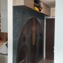 Ściana w mieszkaniu z dekoracyjną, ciemną powłoką imitującą beton z geometrycznym wzorem w odcieniach brązu i złota, nad nią szafka pod sufit, widok z perspektywy wnętrza.