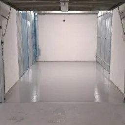 Puste, pomalowane na szaro pomieszczenie magazynowe z metalową konstrukcją sufitu i ścianami z blachy trapezowej, oświetlone pojedynczą żarówką.