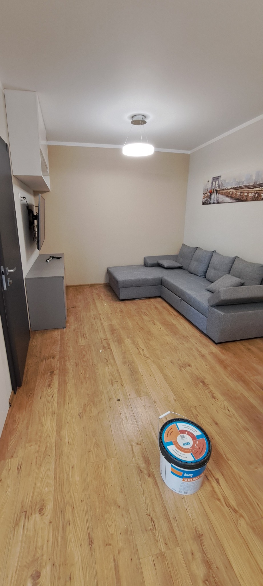 Jasne wnętrze po remoncie: szary narożnik, drewniana podłoga, ściany w odcieniach beżu i wiadro z gładzią Knauf Goldband na podłodze. Minimalistyczny wystrój.