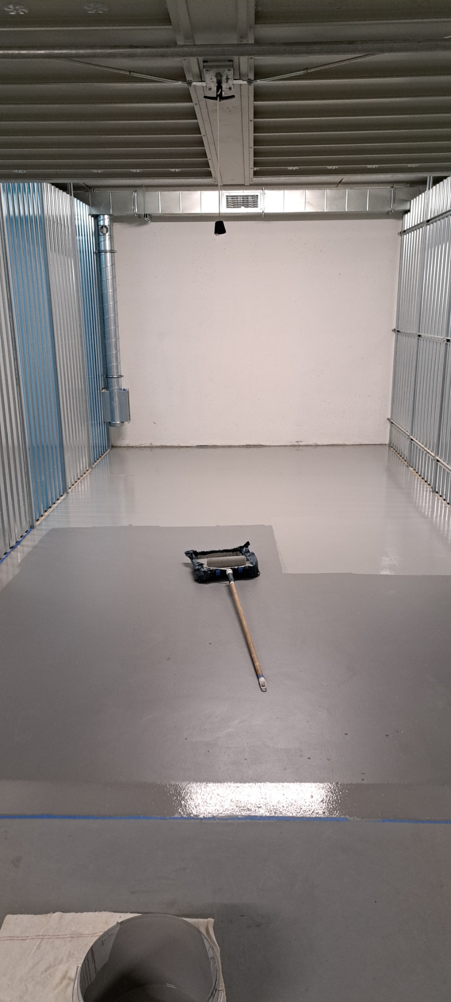 Malowanie podłogi w szarym odcieniu w pomieszczeniu z blachy falistej. Widoczny wałek malarski i wiadro z farbą. Częściowo pomalowana podłoga z niebieską taśmą.