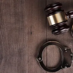 Drewniany stół z sękami, na którym leżą metalowe kajdanki i drewniany młotek sędziowski z pozłacaną podstawą.