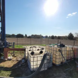 Wiertnica w trakcie pracy na polu, widoczne zbiorniki na wodę i generator, słoneczny dzień.