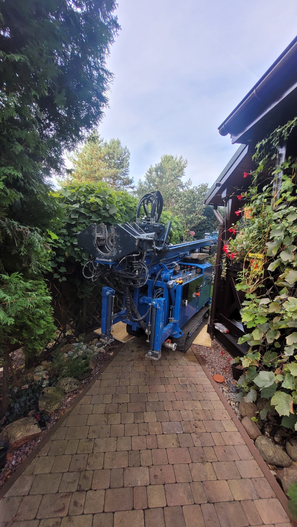 Kompaktowa, niebieska wiertnica gąsienicowa ustawiona na kostce brukowej w ogrodzie, pomiędzy budynkiem a gęstą roślinnością.