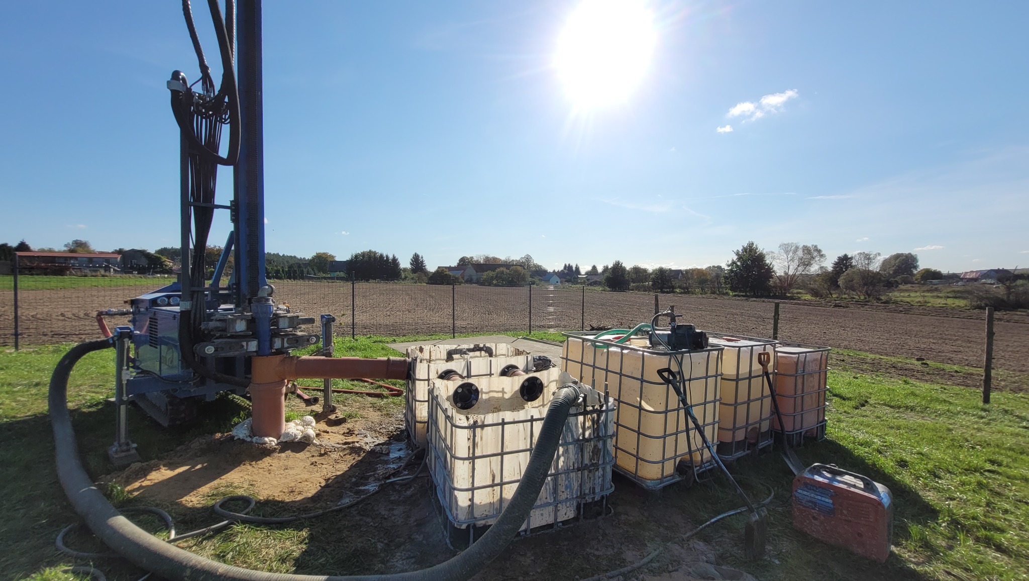 Wiertnica w trakcie pracy na polu, widoczne zbiorniki na wodę i generator, słoneczny dzień.