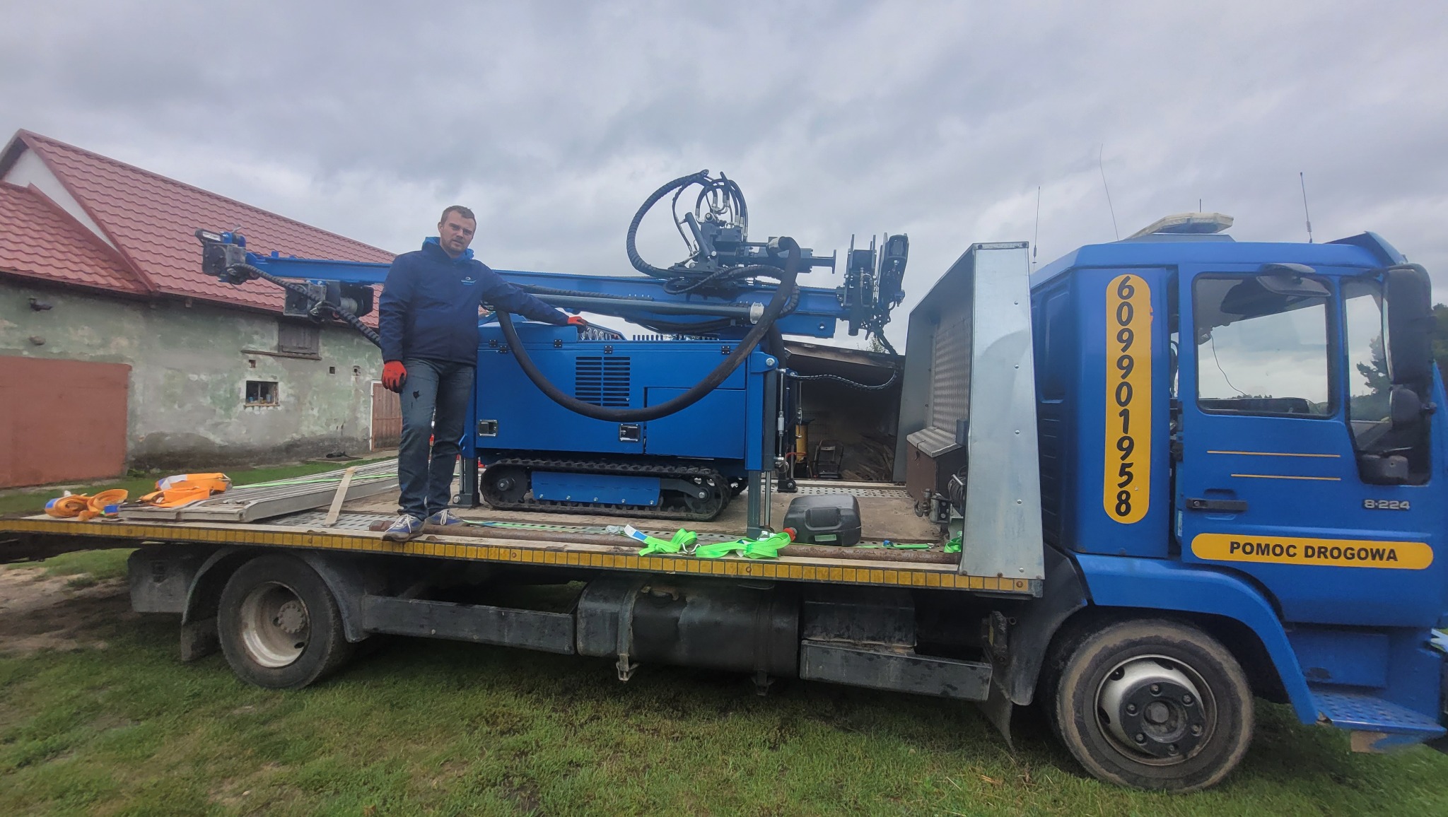 Niebieska wiertnica gąsienicowa na lawecie ciężarówki pomocy drogowej, operator w rękawicach wskazuje na maszynę, w tle budynek gospodarczy z czerwonym dachem.