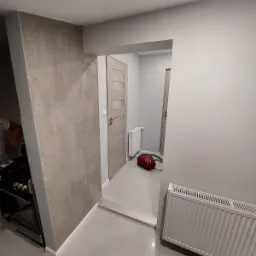 Wnętrze po odświeżeniu: ściana z dekoracyjną fakturą obok białej listwy przypodłogowej, grzejnik na ścianie, drzwi z przeszkleniami i odkurzacz na podłodze.