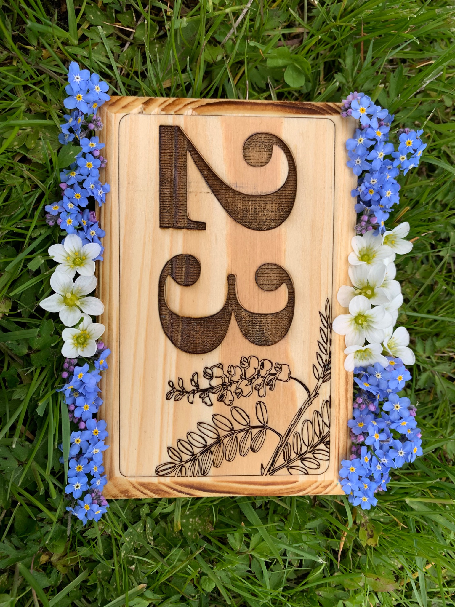 Drewniana tabliczka z wygrawerowanym numerem 23 i motywem roślinnym, otoczona niebieskimi niezapominajkami i białymi kwiatami, leżąca na trawie.
