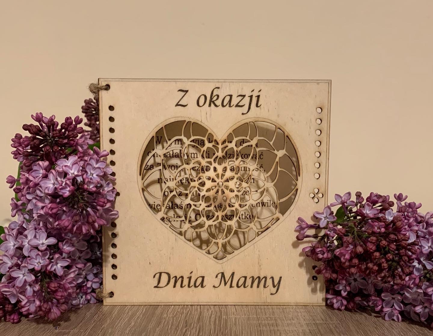 Drewniana kartka okolicznościowa z wyciętym laserowo wzorem serca i napisem 'Z okazji Dnia Mamy', ozdobiona gałązkami bzu po bokach.