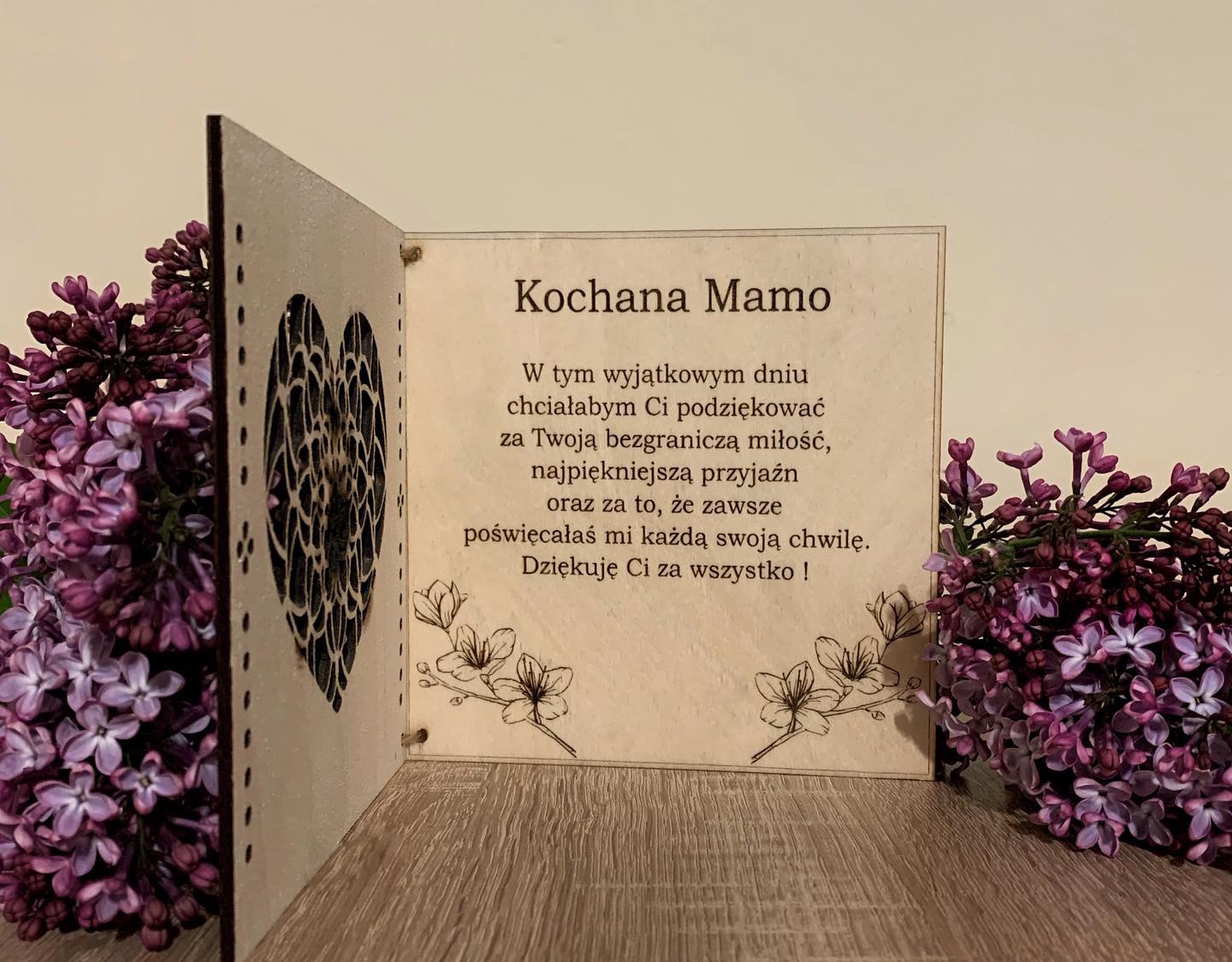 Drewniana kartka z wyciętym sercem i dedykacją 'Kochana Mamo', udekorowana gałązkami kwitnącego bzu w odcieniach fioletu.