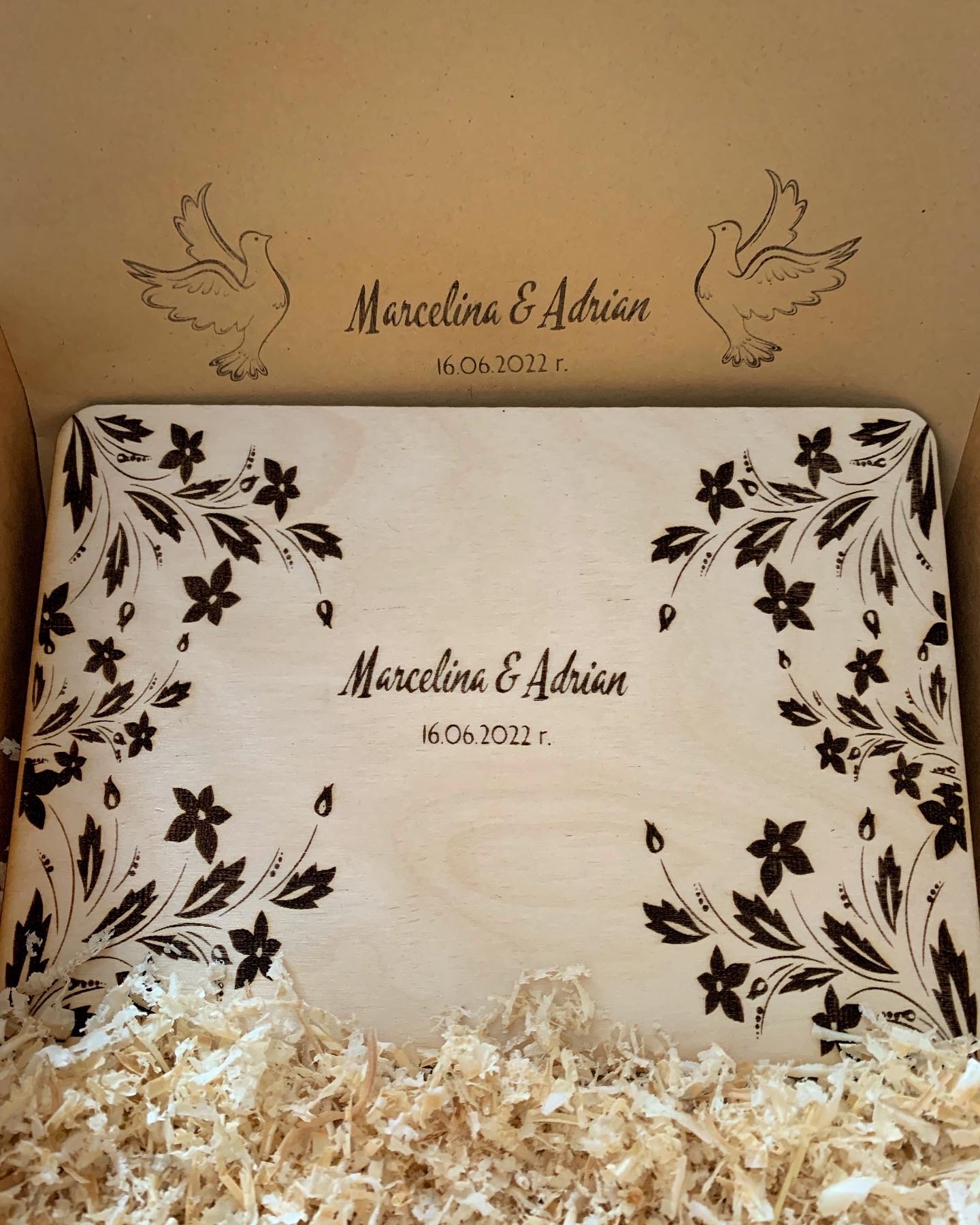 Drewniane pudełko z wygrawerowanymi imionami Marcelina i Adrian oraz datą 16.06.2022, ozdobione motywem kwiatowym i gołębiami, wypełnione wiórkami drzewnymi.