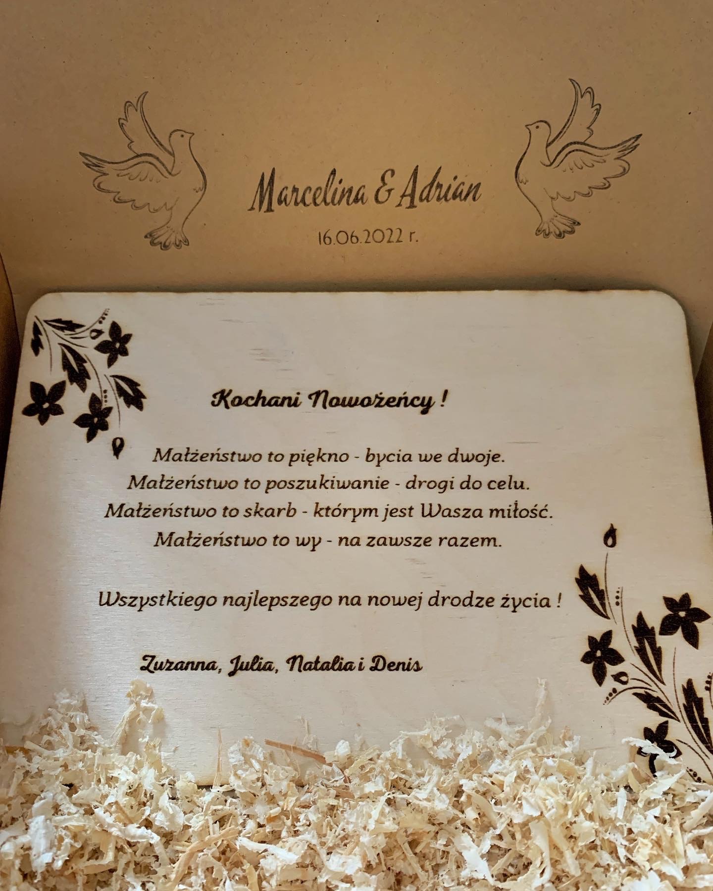Drewniane pudełko z wygrawerowaną dedykacją dla Marceliny i Adriana z okazji ślubu 16.06.2022, ozdobione rysunkami gołębi i kwiatów, wypełnione wiórkami.