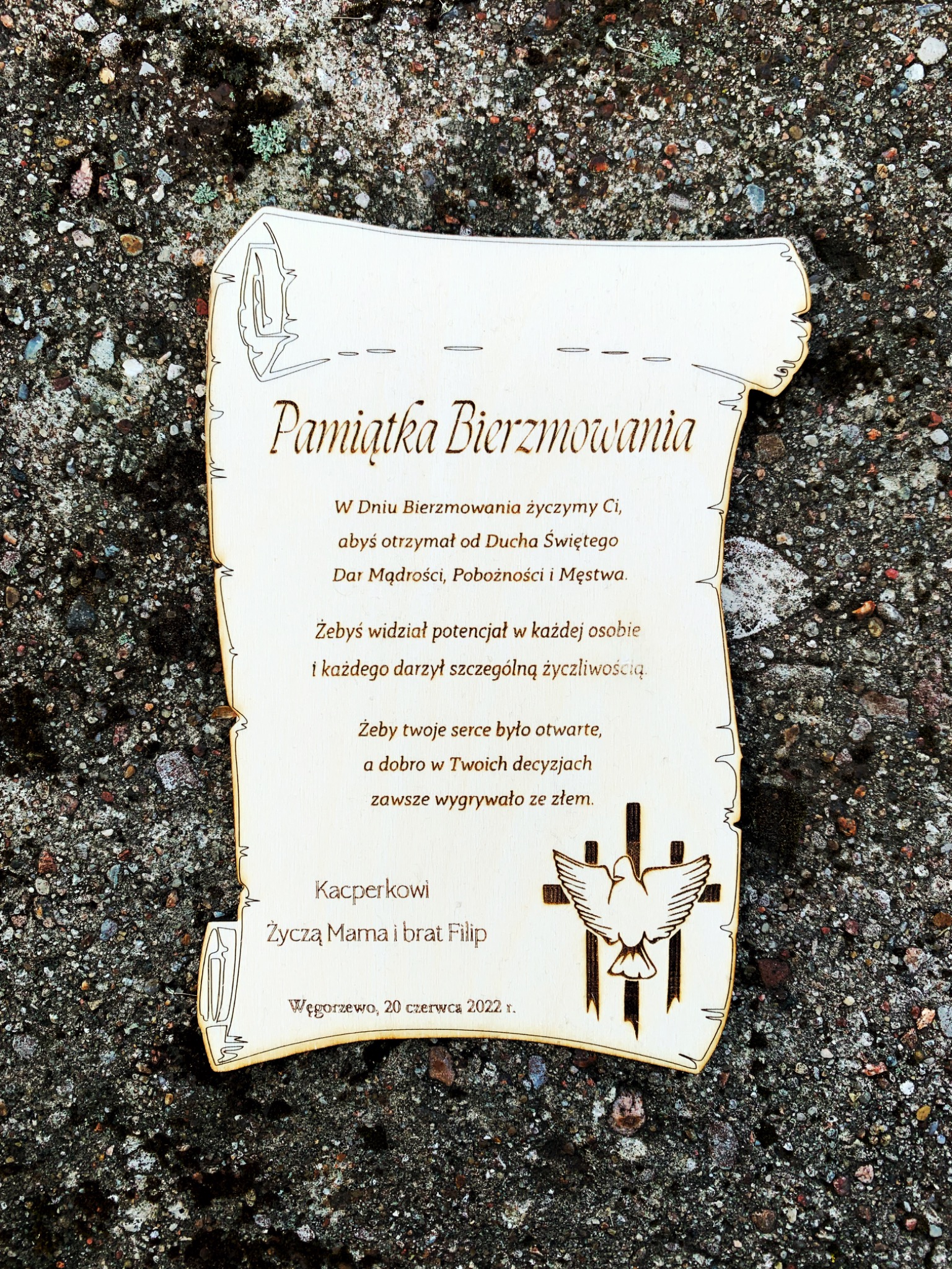 Drewniana pamiątka bierzmowania w kształcie zwoju, z wygrawerowanym tekstem życzeń i symbolem gołębicy, leżąca na szorstkim, kamiennym podłożu.