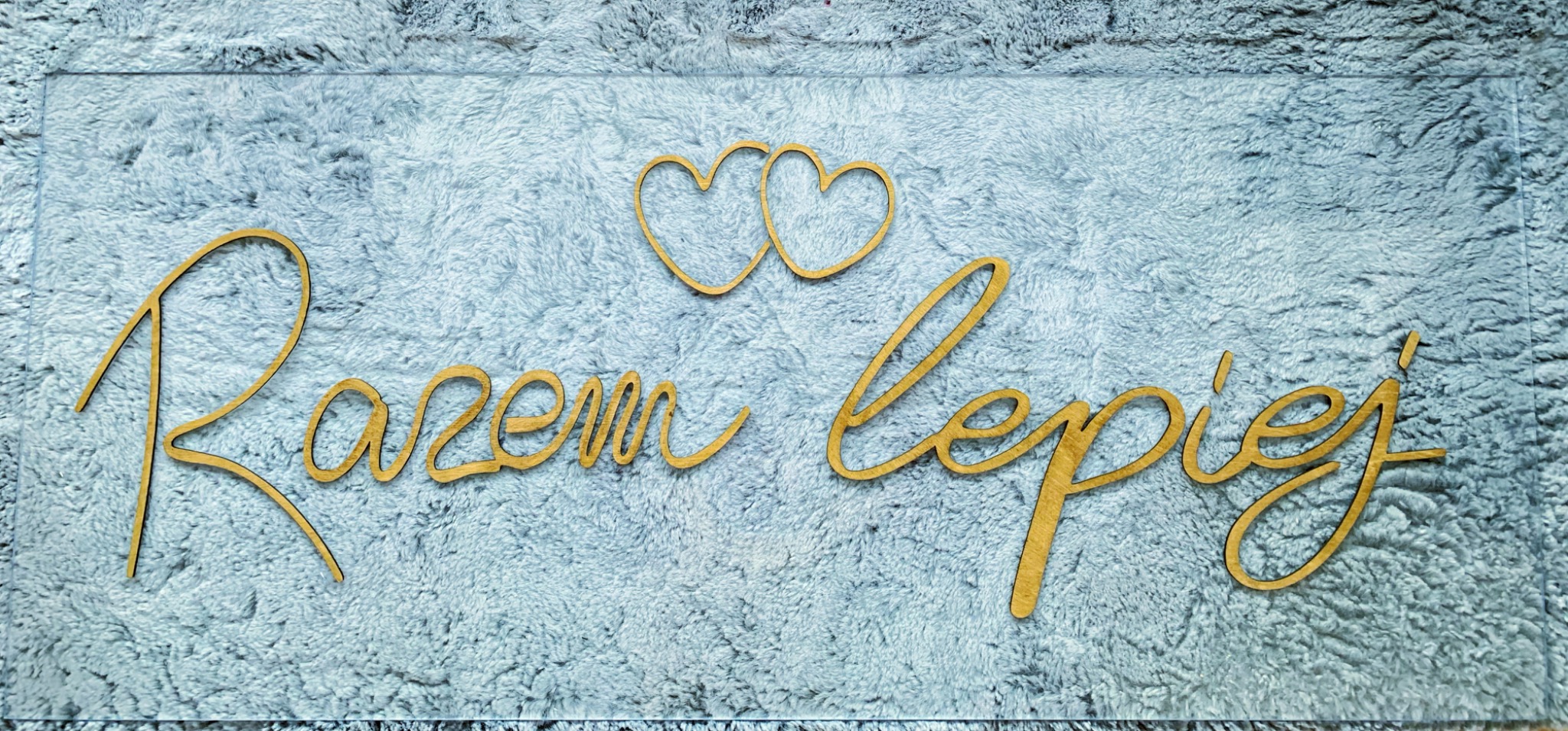 Dekoracja ślubna z napisem 'Razem Lepiej' i dwoma sercami, wykonana z drewnianych elementów na tle szarej, puszystej tkaniny, umieszczona na przezroczystej płycie.