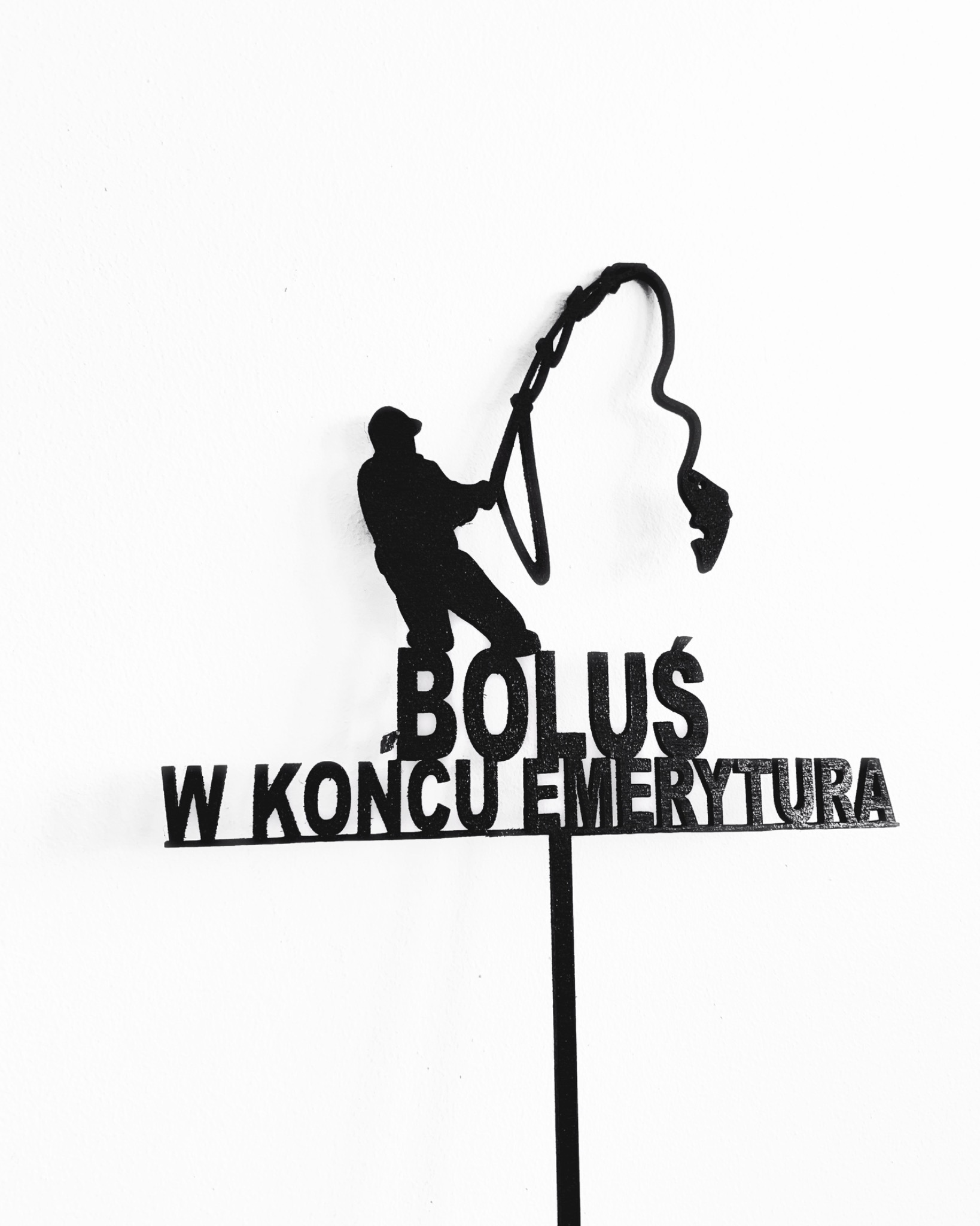 Czarna, brokatowa dekoracja tortu przedstawiająca sylwetkę wędkarza ciągnącego rybę i napis 'Boluś. W końcu emerytura', umieszczona na białym tle.