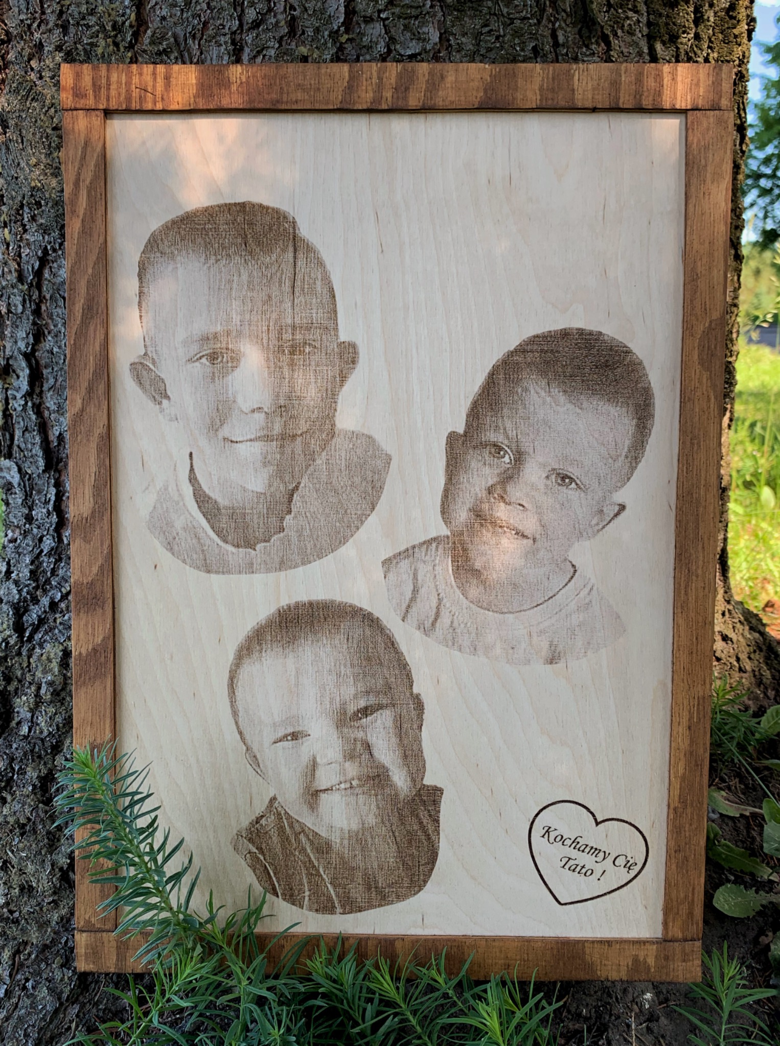 Drewniana tablica z wygrawerowanymi portretami trzech chłopców i napisem 'Kochamy Cię Tato!' w kształcie serca, wykonana w Węgorzewie, oparta o korę drzewa.