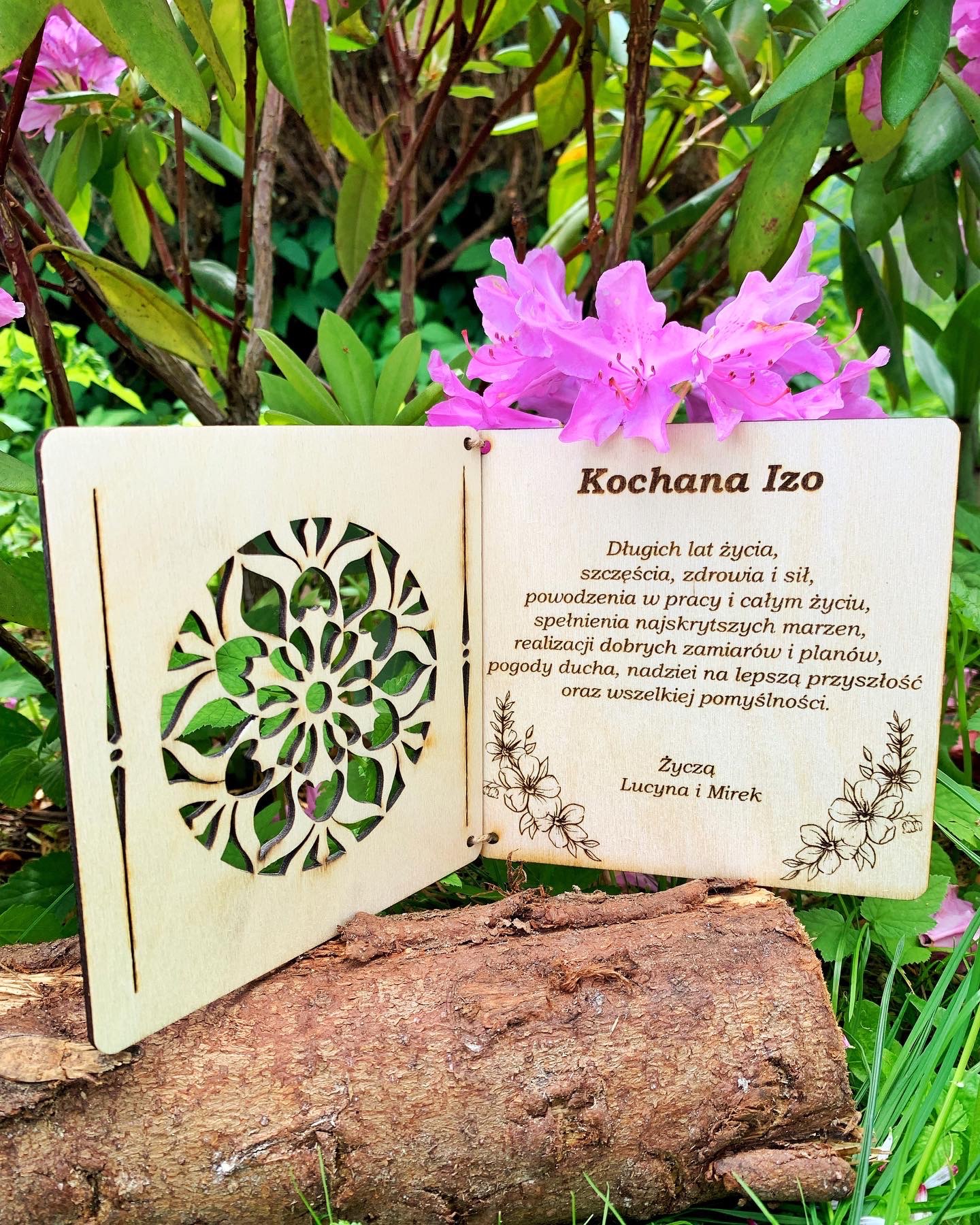 Drewniana, laserowo wycinana kartka z życzeniami dla Izy, z motywem kwiatowym i ażurowym wzorem, umieszczona na kawałku drewna na tle zieleni i różowych kwiatów.